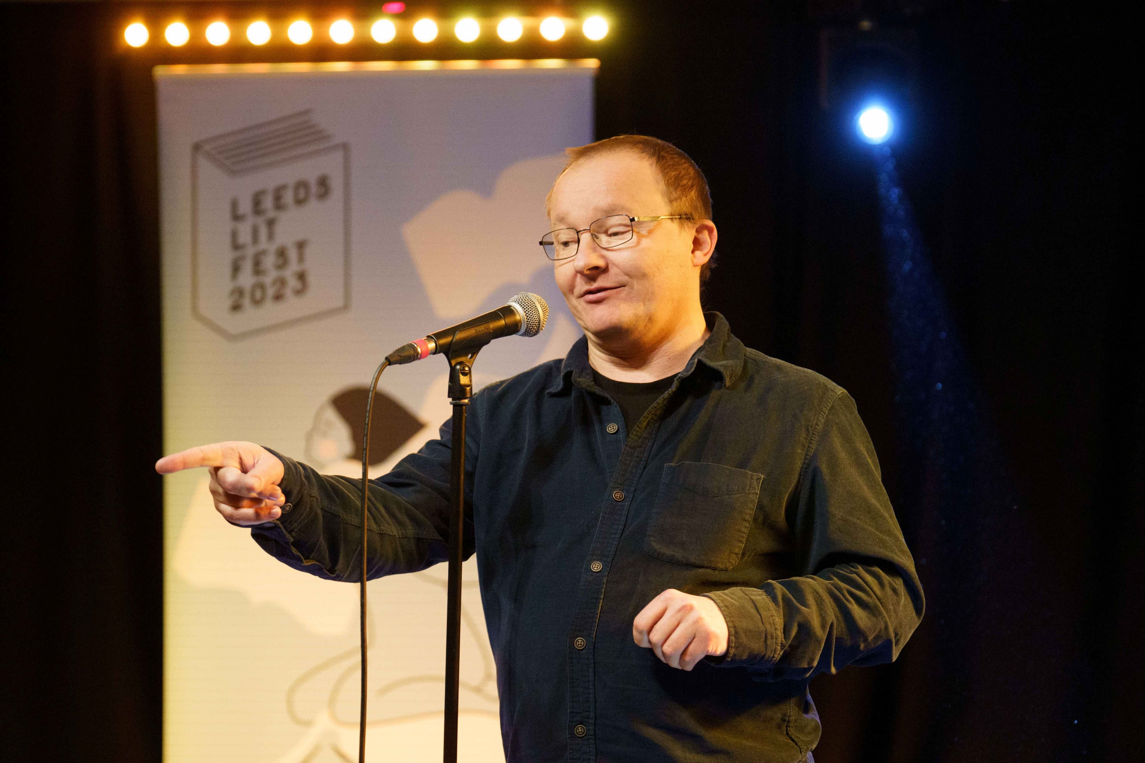A speaker gestures passionately at a microphone during Leeds Lit Fest, with a colorful backdrop and stage lights.
