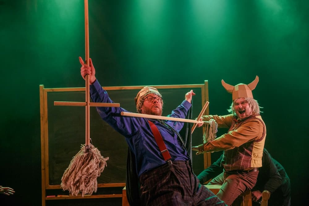 Two comedic performers in vibrant costumes act out a playful scene on stage, illuminated by green stage lights.