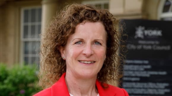 A woman with curly hair in a red blazer stands in front of the City of York Council building, smiling warmly.