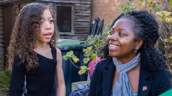 A smiling woman interacts with a young girl outdoors, surrounded by greenery and rustic buildings in York.