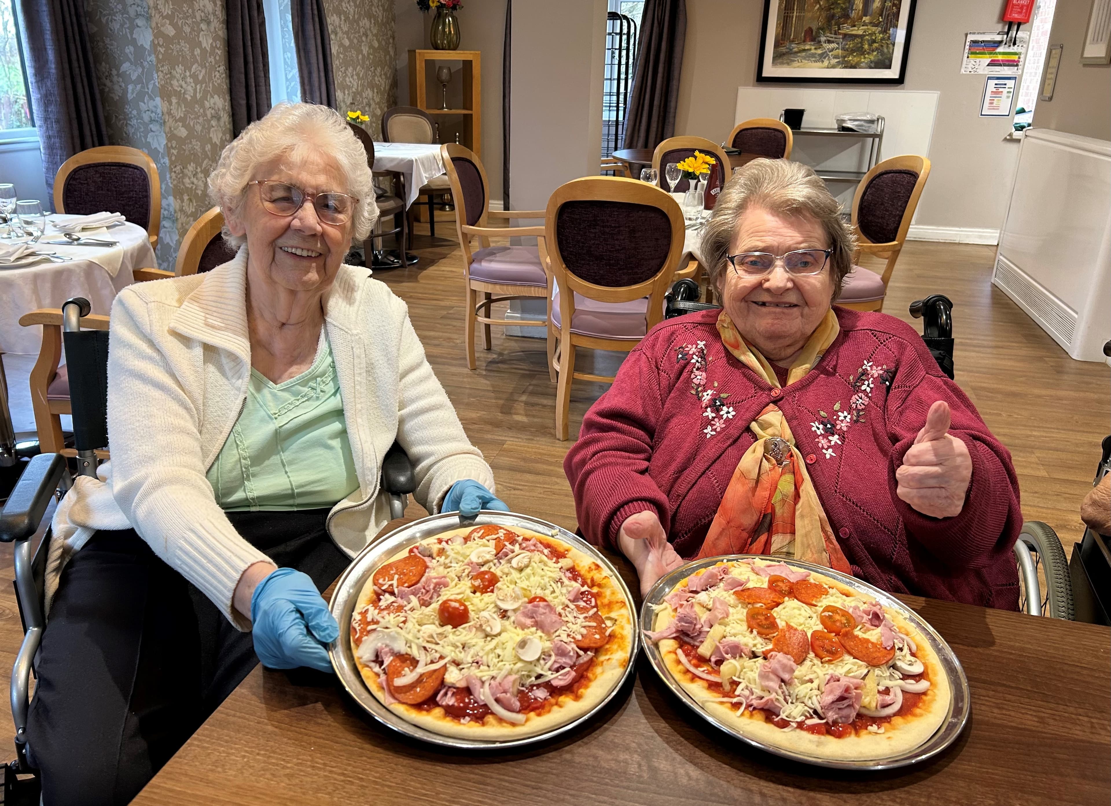 Two elderly women in a dining room holding homemade pizzas