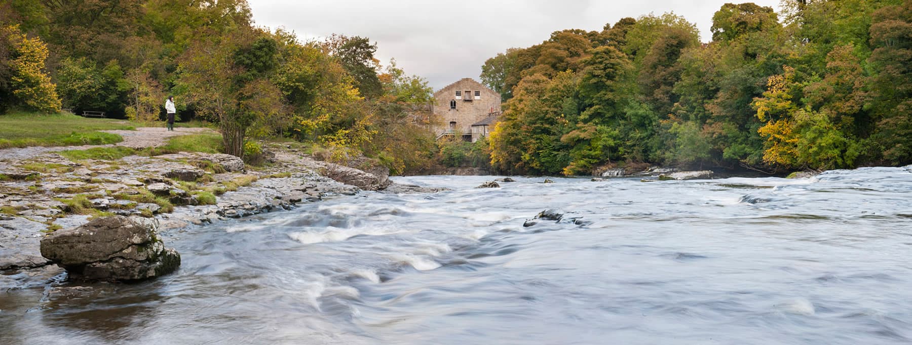 Rushing river flanked by rocky banks and lush trees, with a stone building visible in the background.