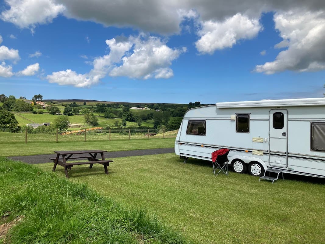 Caravan parked on green grass with picnic table, surrounded by rolling hills and a blue sky with fluffy clouds.