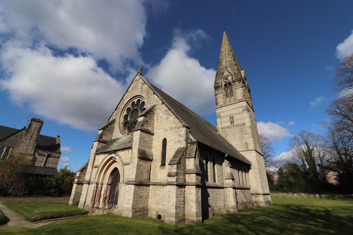 Victorian church with a tall spire, large rose window, and stone facade, surrounded by green grass and trees.