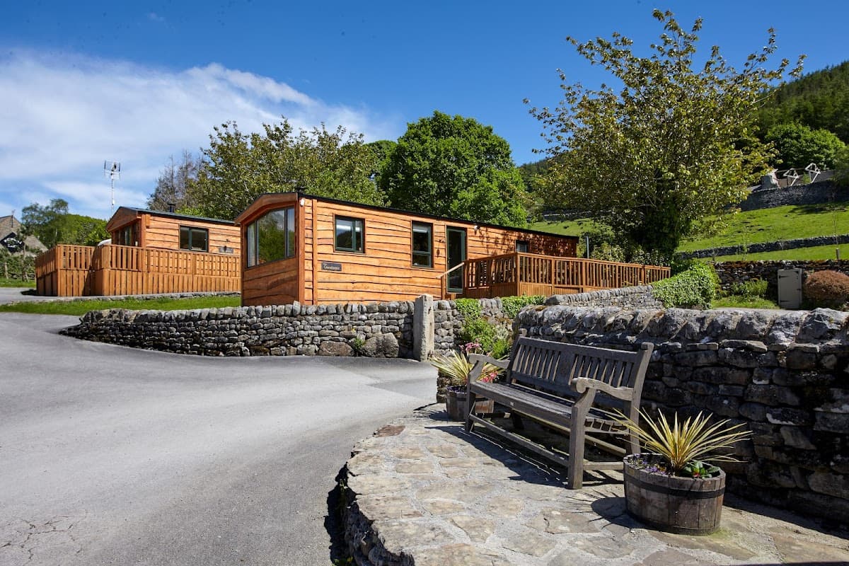 Wooden lodges with decking surrounded by greenery, stone walls, and a bench in a scenic Yorkshire park.