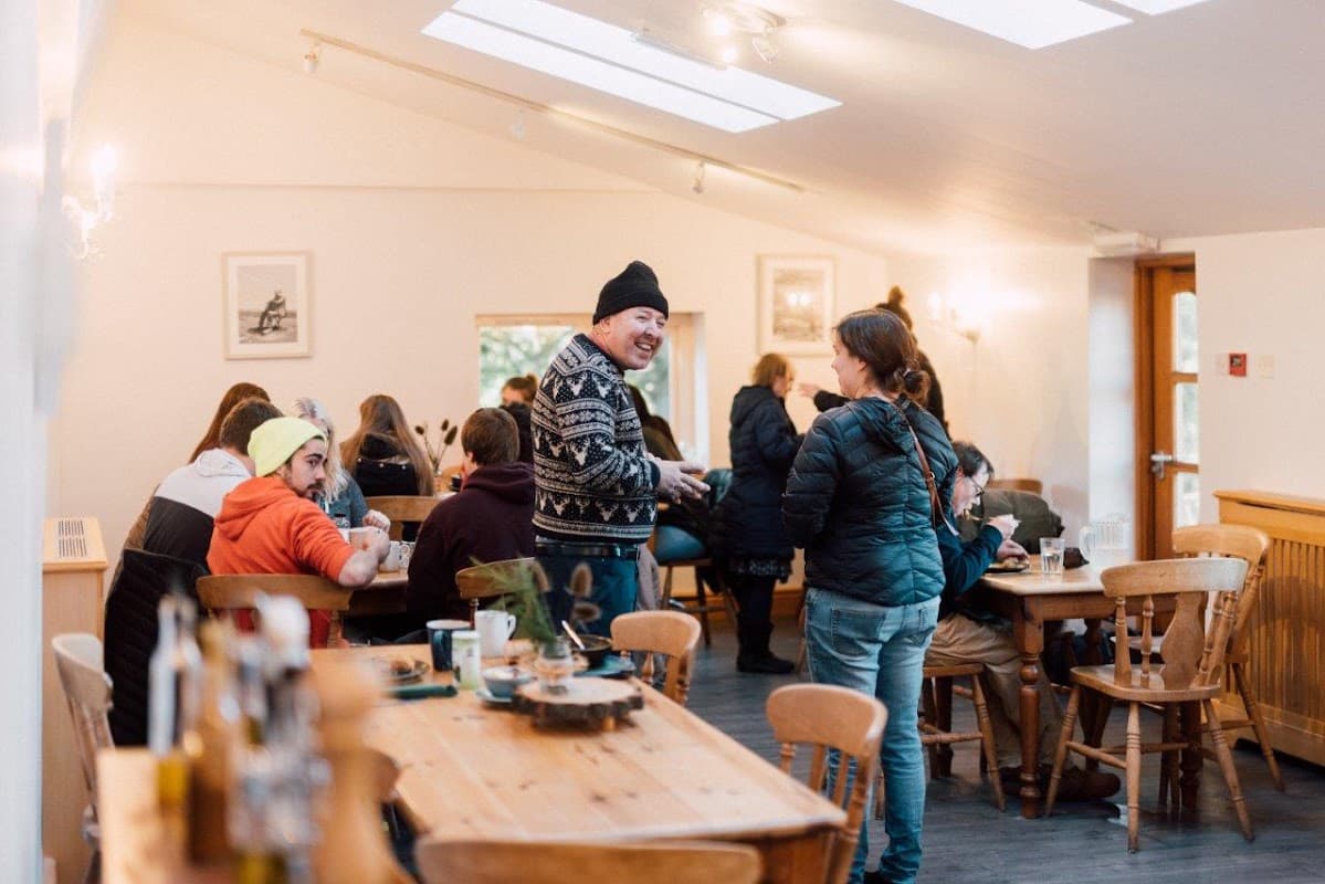 Cozy cafe interior with patrons seated at wooden tables, chatting, and a staff member interacting with a customer.