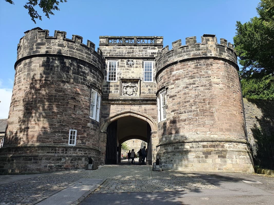 Historic stone gatehouse with towers, surrounded by greenery and a cobbled path, leading into a park area.