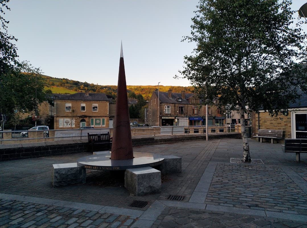 St. Michaels Square features a tall metal sculpture, stone benches, and buildings with a hillside backdrop in Chiserley, Yorkshire.