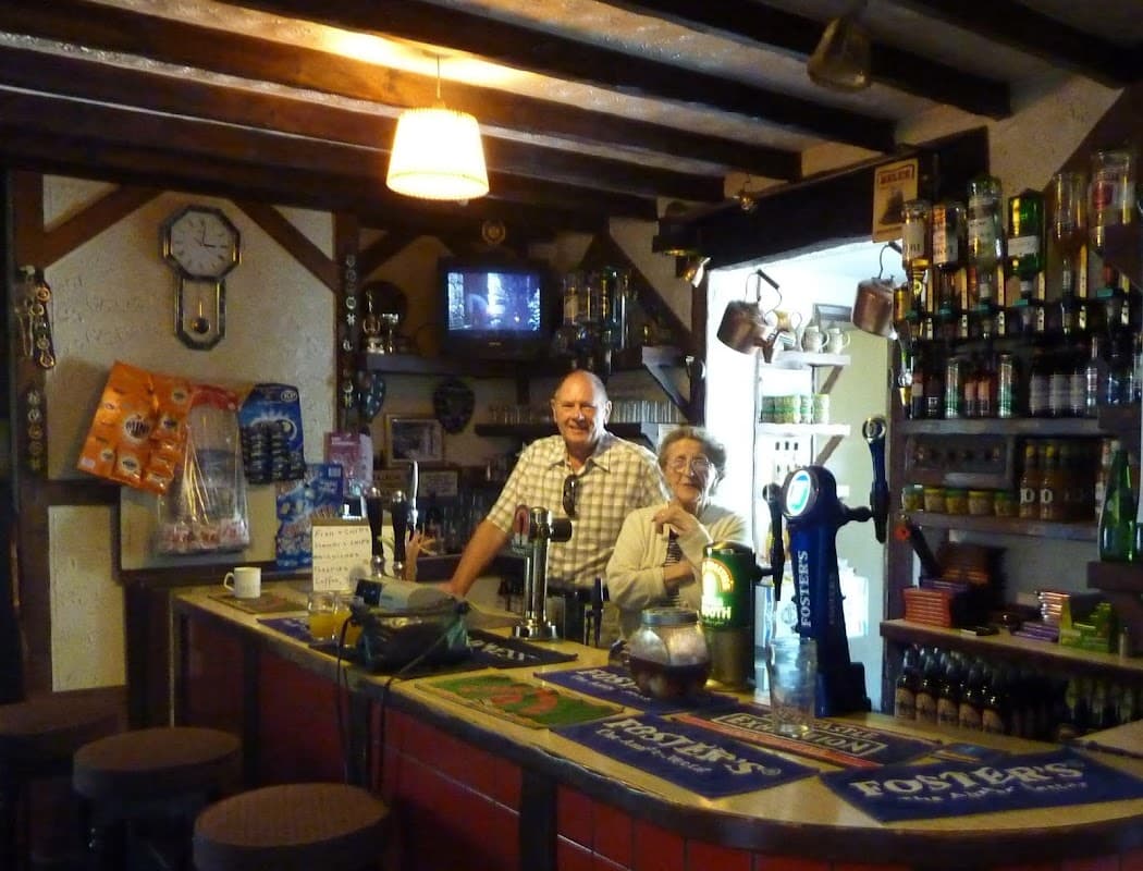 Bar interior featuring a counter, two people smiling, shelves of drinks, and a vintage clock on the wall.