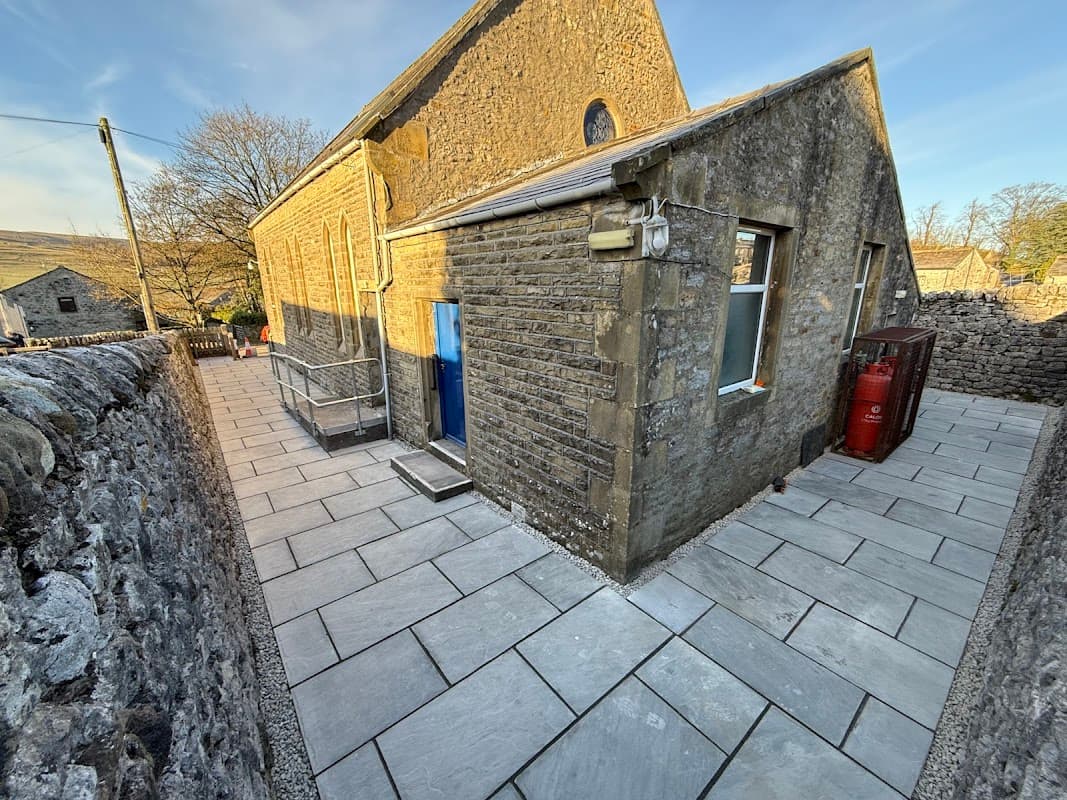 Stone building with a blue door, large windows, and a paved pathway, surrounded by a dry stone wall and trees.