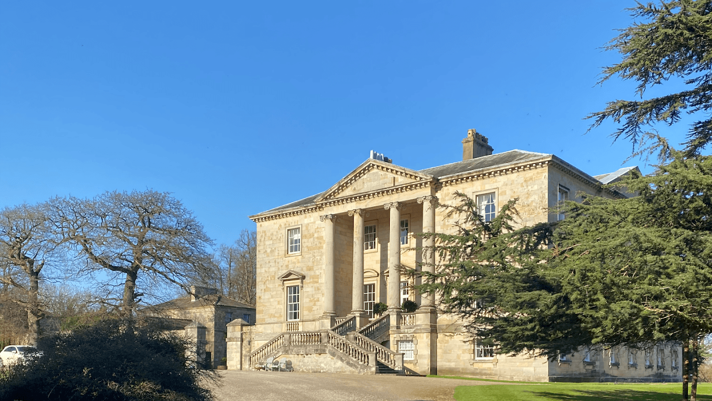 Historic stone building with columns, surrounded by lush gardens and trees under a clear blue sky.