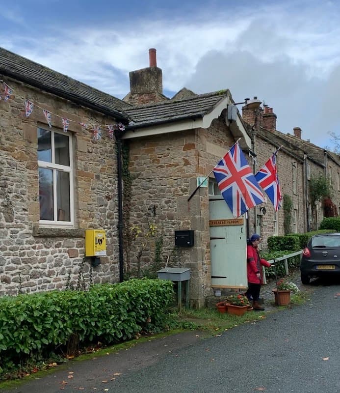 Stone building with Union Jack flags, a woman standing outside, and a parked car in a quaint Yorkshire village.