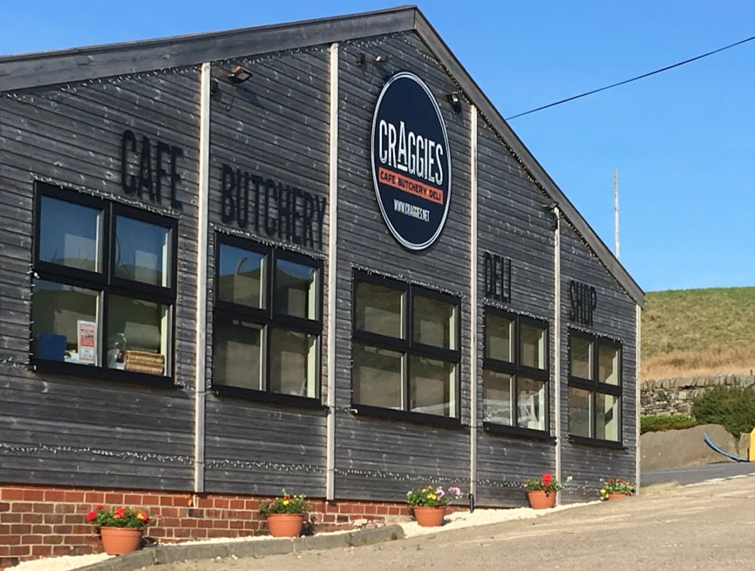 Wooden building with large windows, sign reading "Craggies Cafe Butchery Deli Shop," and colorful flower pots outside.