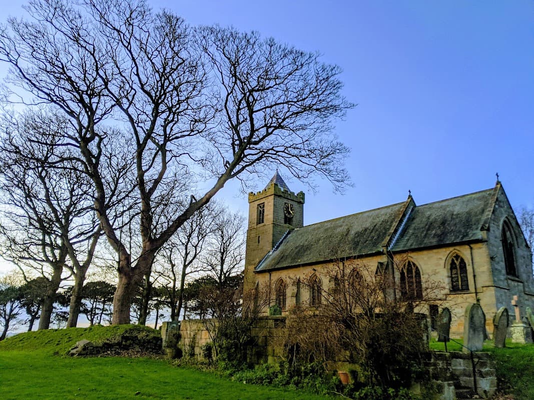 All Saints Church with a tall tree nearby, set against a clear blue sky and grassy grounds.