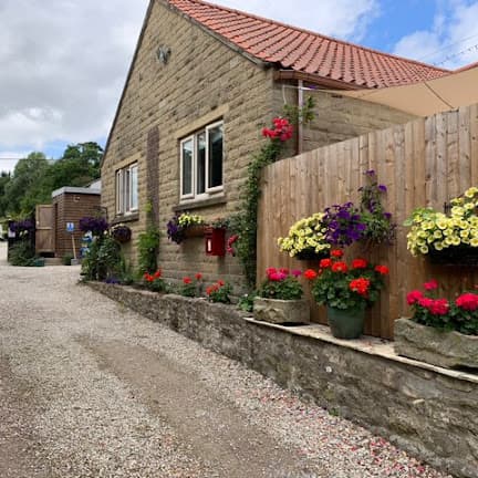 Charming stone building with vibrant flower pots and hanging baskets along a gravel pathway in a scenic campsite.