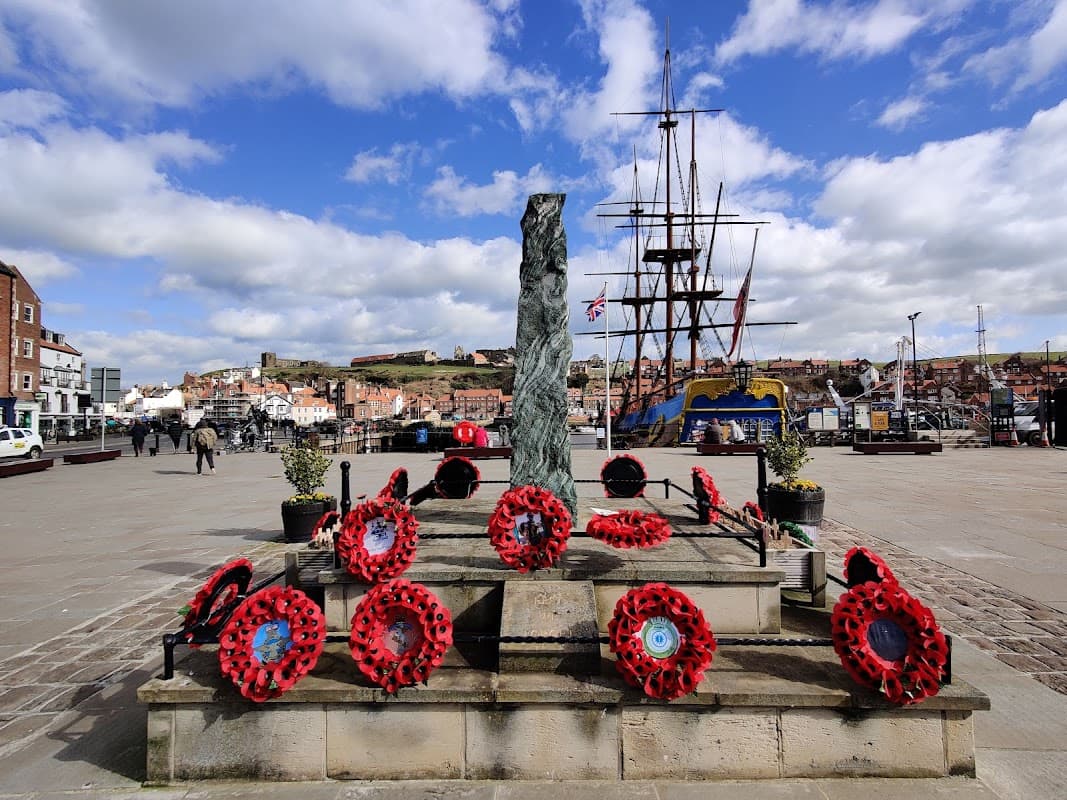 War memorial adorned with red poppies, overlooking a harbor with ships and a backdrop of Whitby's buildings and hills.