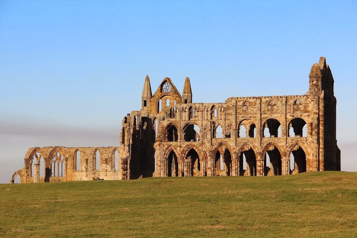 Ruins of Whitby Abbey stand against a clear blue sky, surrounded by green grass in Yorkshire.
