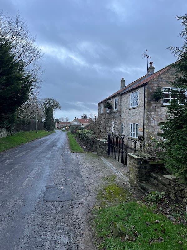 Stone cottage along a quiet road, surrounded by greenery and trees under a cloudy sky in Fadmoor, Yorkshire.