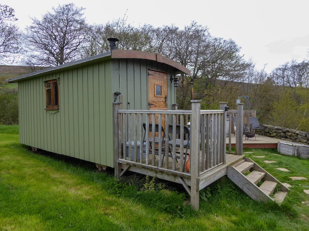 Green cabin with wooden deck, steps, and outdoor seating surrounded by grassy landscape and trees in Fryup, Yorkshire.