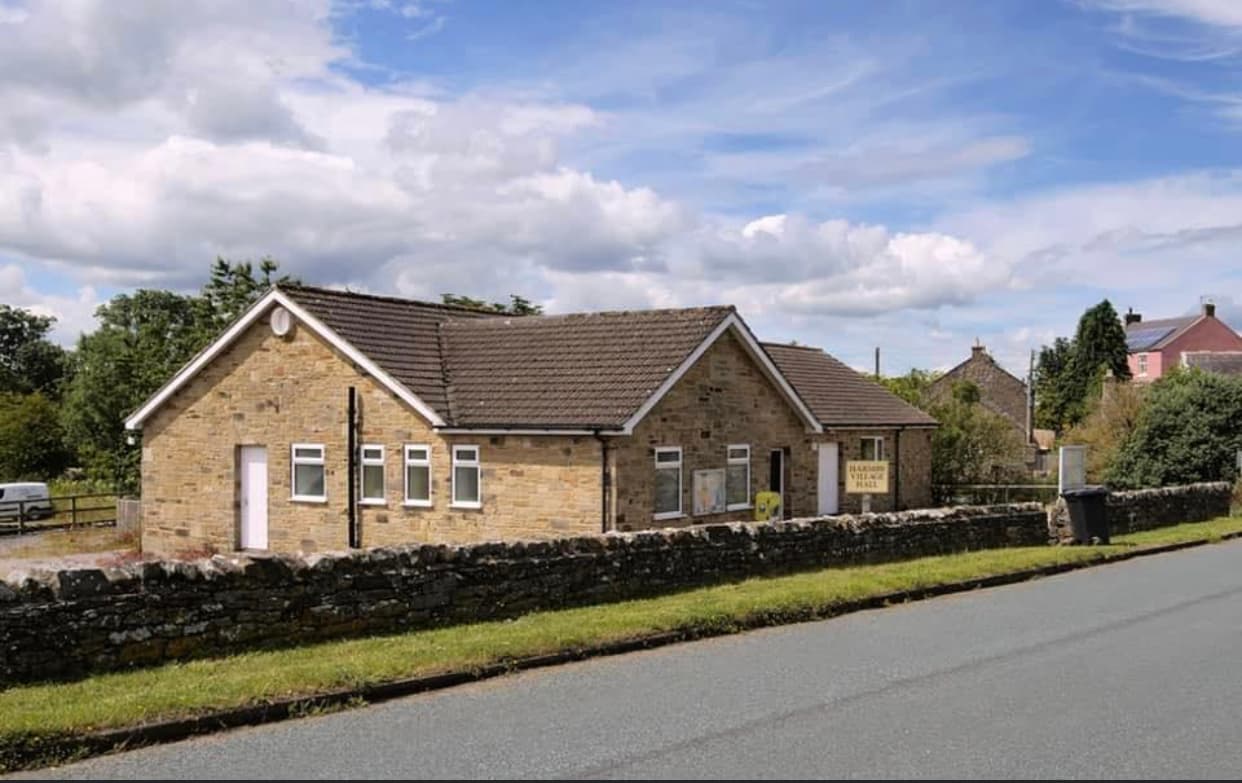Stone building with a sloped roof, surrounded by greenery and a stone wall, along a quiet road in Harmby, Yorkshire.