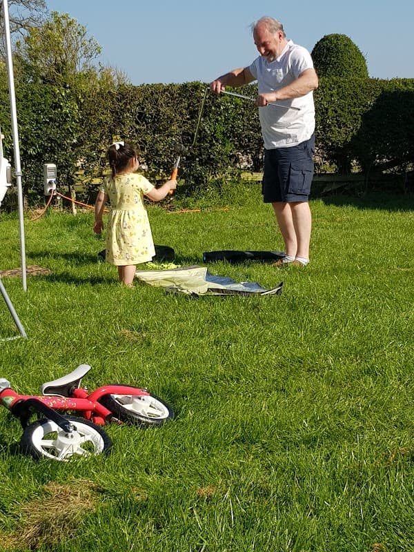 A man and a child play with bubbles in a grassy area, with a tricycle nearby and hedges in the background.