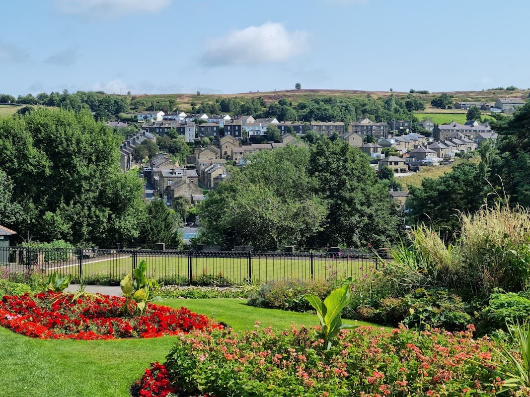 Lush green park with vibrant flowerbeds, overlooking a charming village and rolling hills under a clear blue sky.