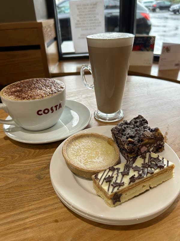 Cappuccino, latte, and three desserts: a tart, brownie, and chocolate slice on a wooden table at Costa Coffee.