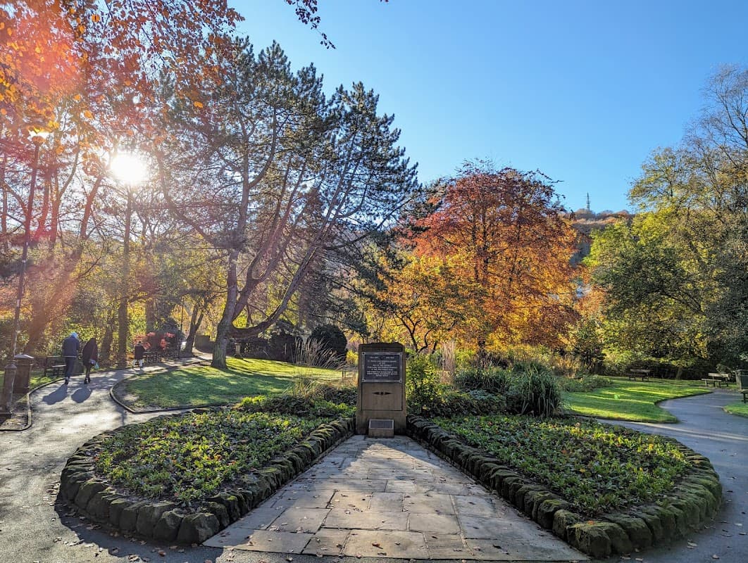 A serene memorial garden with vibrant autumn foliage, stone pathways, and a central plaque under a bright blue sky.