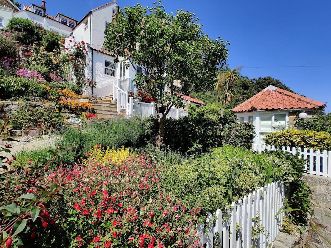 Lush garden with colorful flowers, a white picket fence, and a charming gazebo under a clear blue sky.