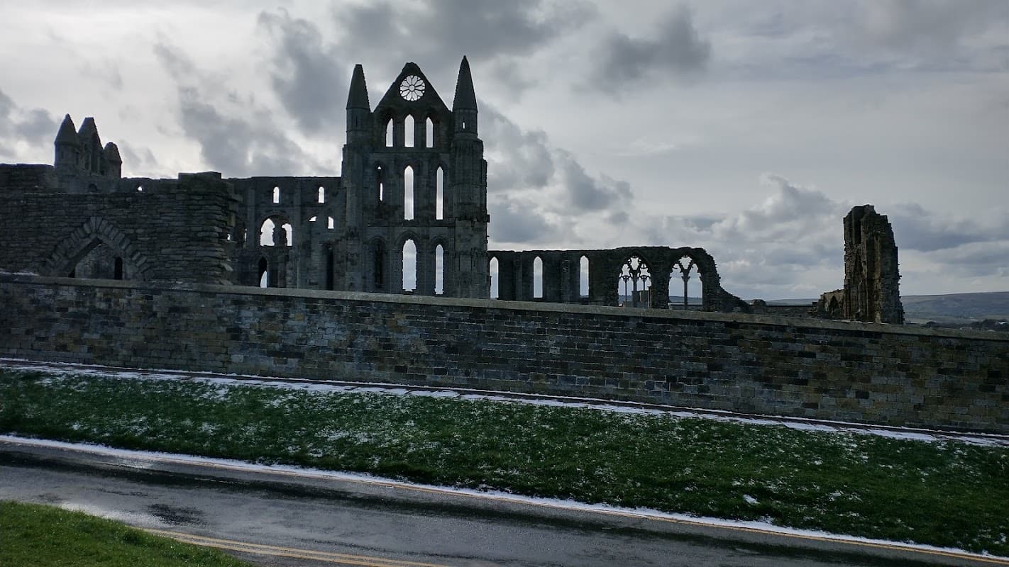 Ruins of a historic abbey with tall arches and windows, set against a cloudy sky in a grassy landscape.