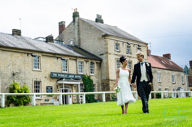 Couple walking hand-in-hand on green lawn in front of the Worsley Arms Hotel, a charming stone building.