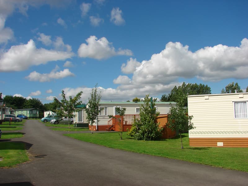 Caravan park with neatly arranged mobile homes, green lawns, trees, and a blue sky dotted with fluffy clouds.