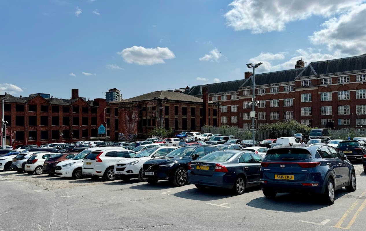 Busy car park filled with parked vehicles, surrounded by brick buildings and blue sky with scattered clouds.