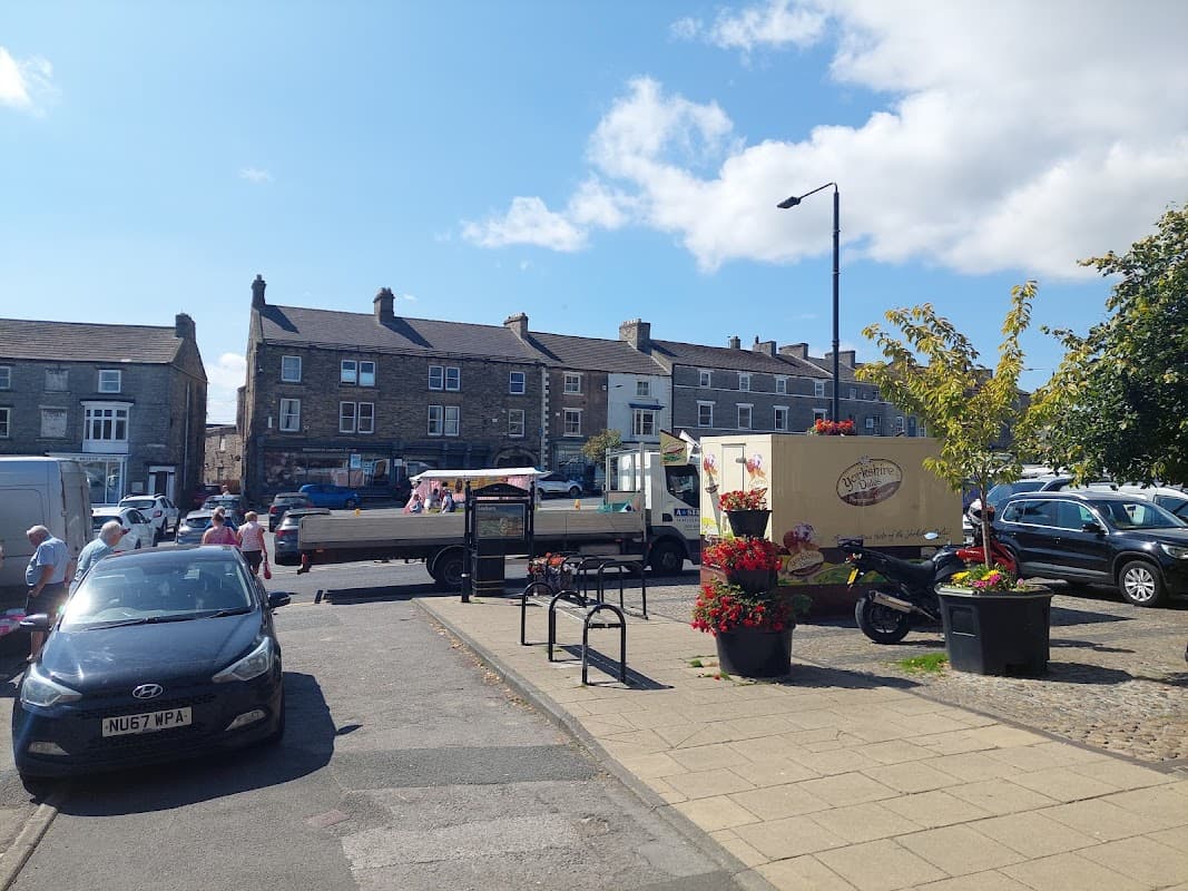 Leyburn Long Stay Car Park with vehicles, shops, and people enjoying a sunny day in Yorkshire.