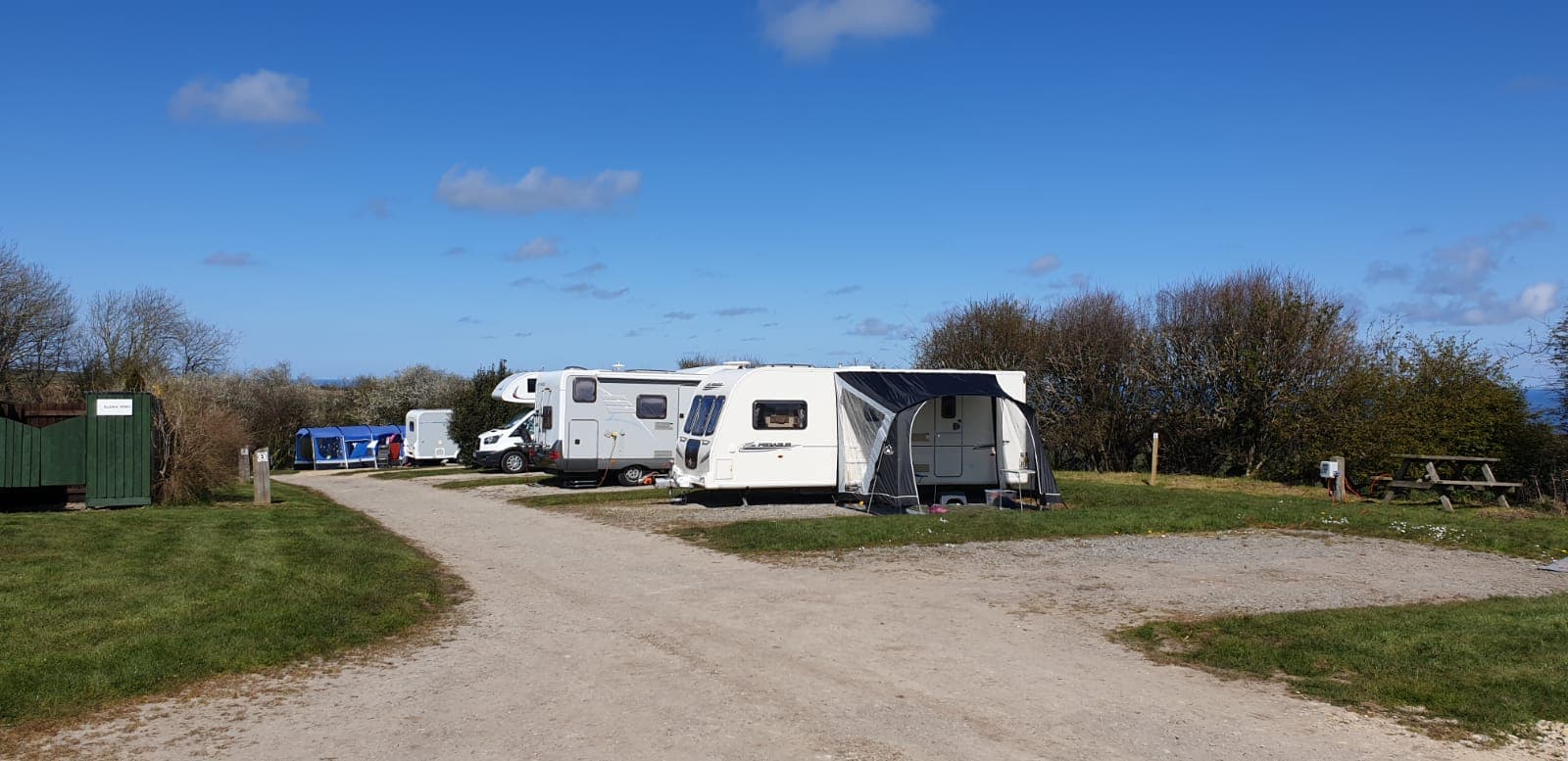 Caravans parked along a gravel path in a grassy area, with blue skies and scattered clouds in the background.