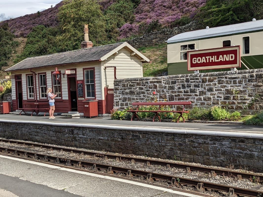 Levisham Station with a quaint building, picnic table, and a sign for Goathland amidst purple heather-covered hills.
