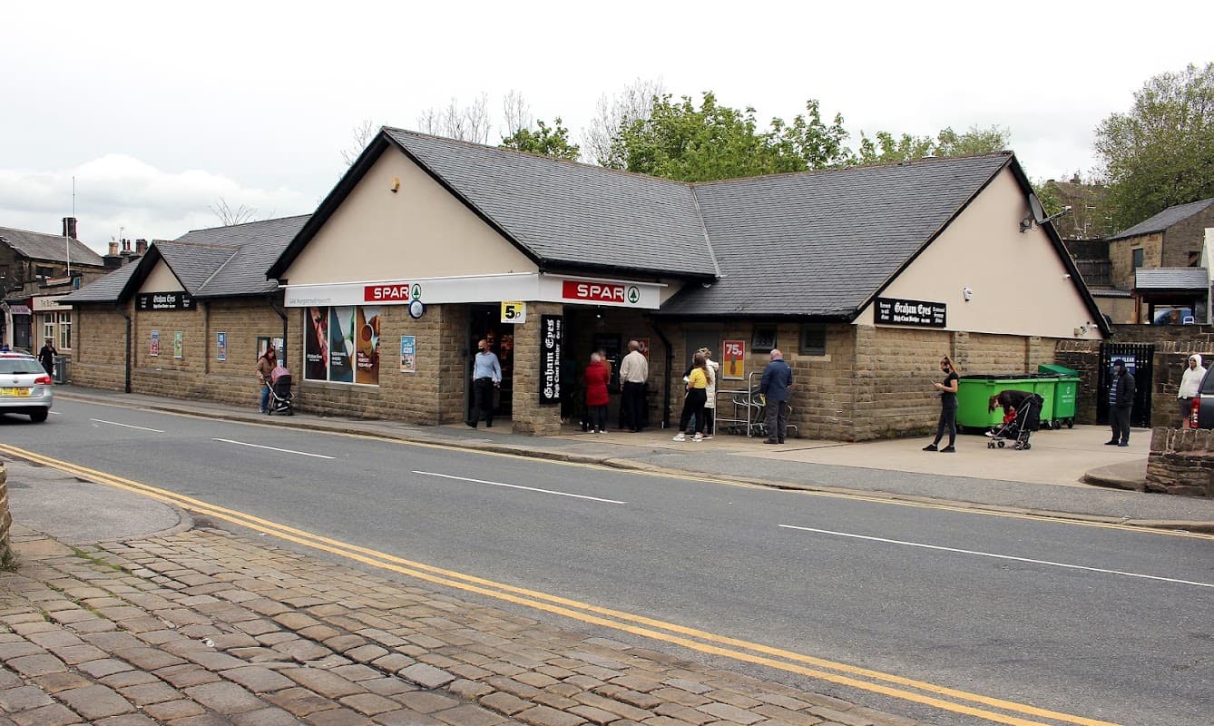 SPAR grocery store in Oxenhope, Yorkshire, with people outside, stone facade, and a green recycling bin nearby.