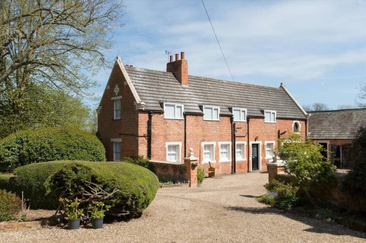 Brick building with multiple windows, surrounded by lush greenery and a gravel pathway in a serene park setting.