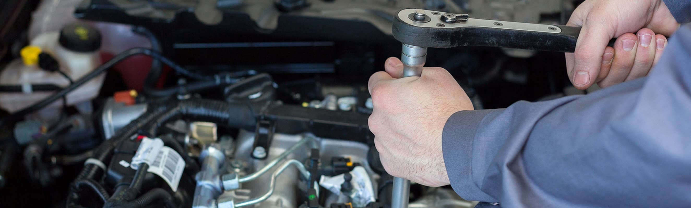 Hands using a ratchet tool on an engine, with various automotive components visible in the background.