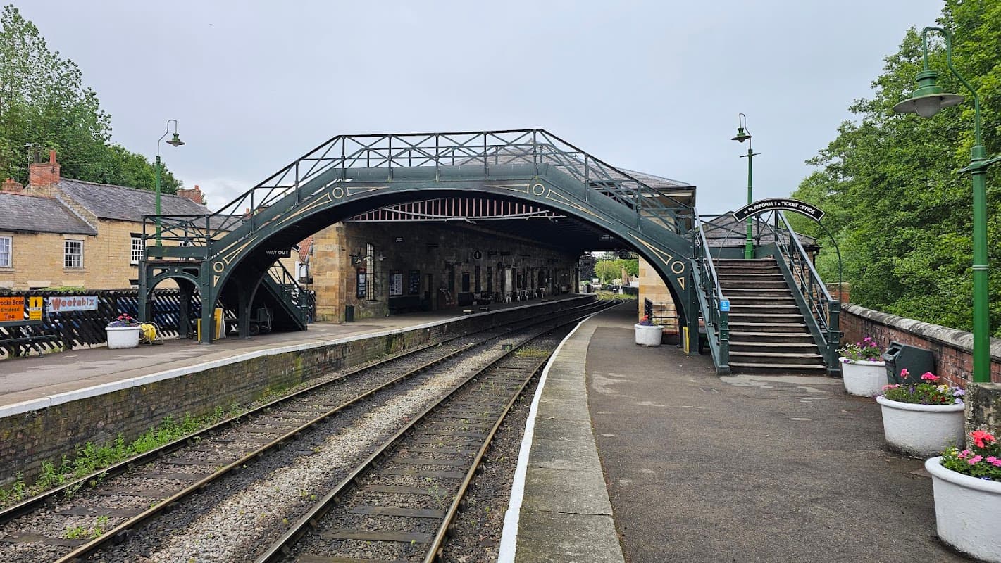 Victorian-style railway station platform with an arched bridge, waiting area, and flower pots along the tracks.