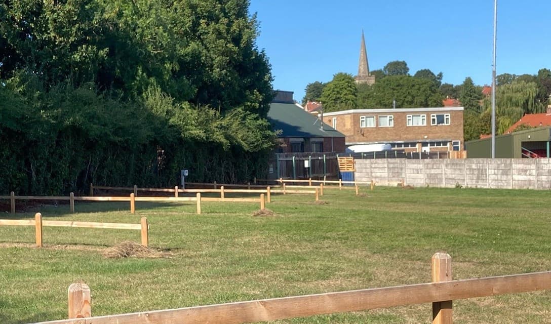 Green field with wooden fences, buildings in the background, and a church spire visible against a clear blue sky.