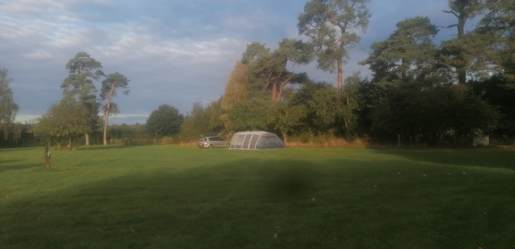 A spacious grassy campsite with a tent and a parked car, surrounded by trees under a partly cloudy sky.