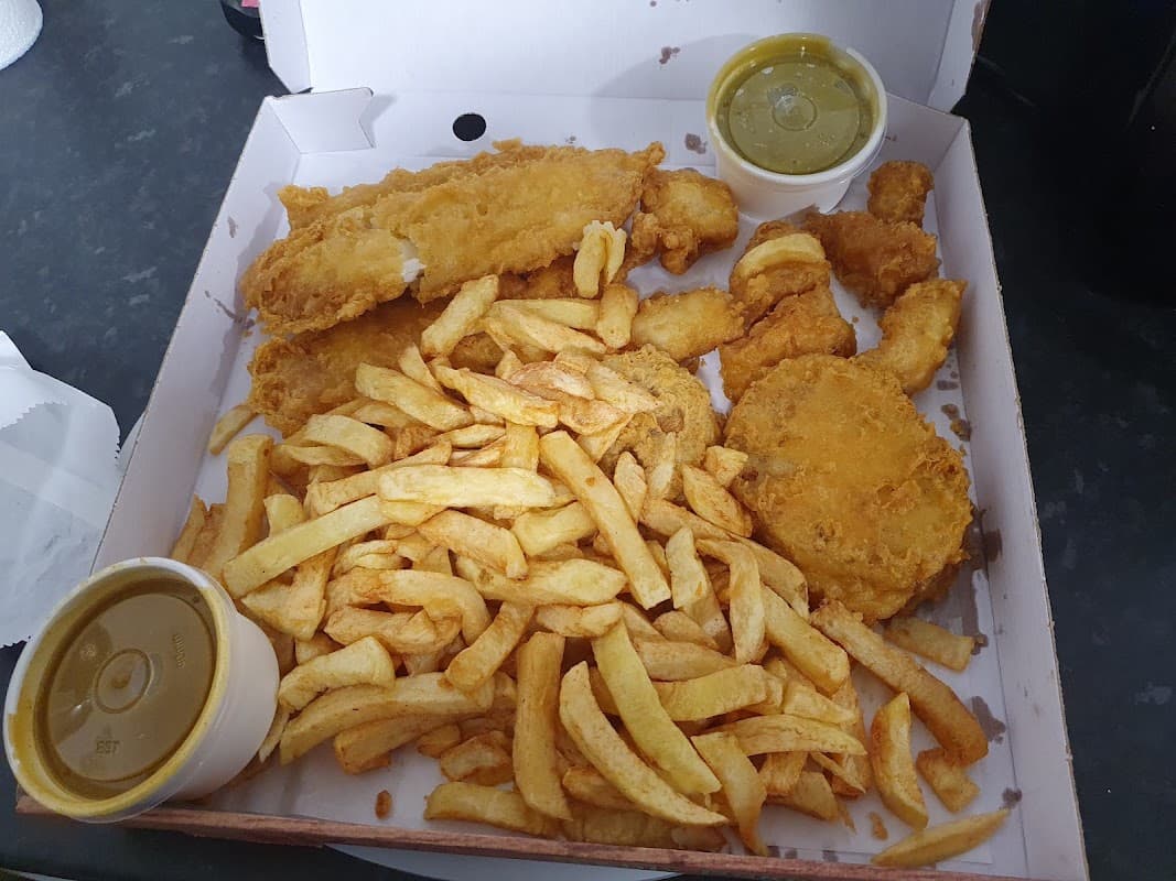 A takeaway box filled with golden fried fish, chicken pieces, and a generous portion of chips, accompanied by two sauces.
