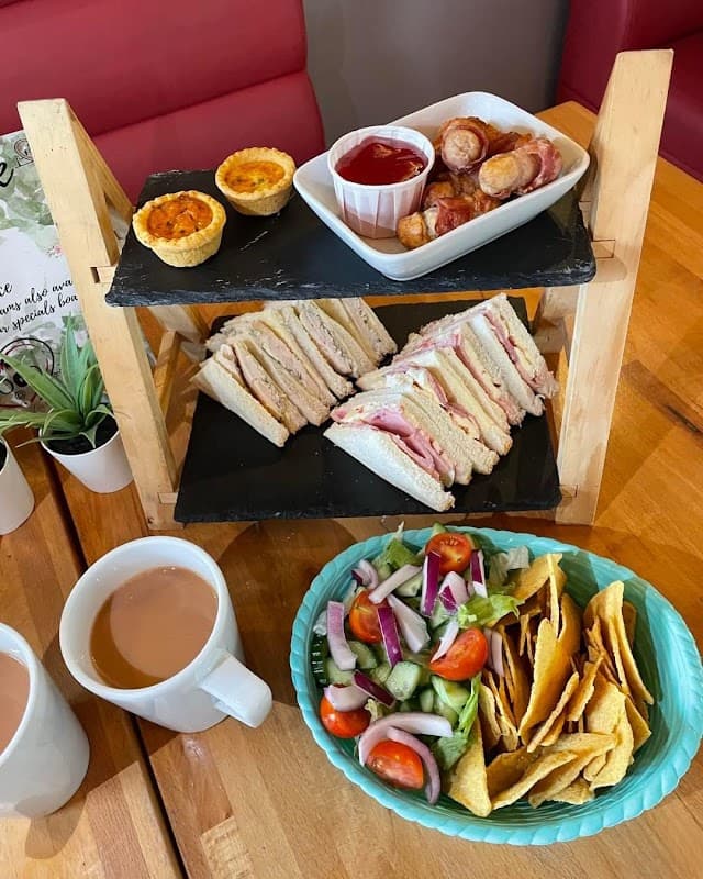 A wooden display holds sandwiches, pastries, and a bowl of sausages, with a side of salad and a cup of tea on the table.