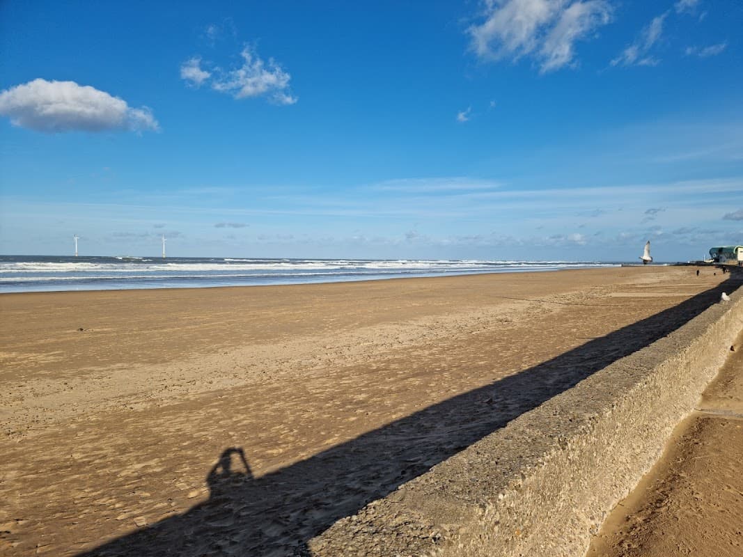 A sandy beach with gentle waves, distant wind turbines, and a clear blue sky with scattered clouds.
