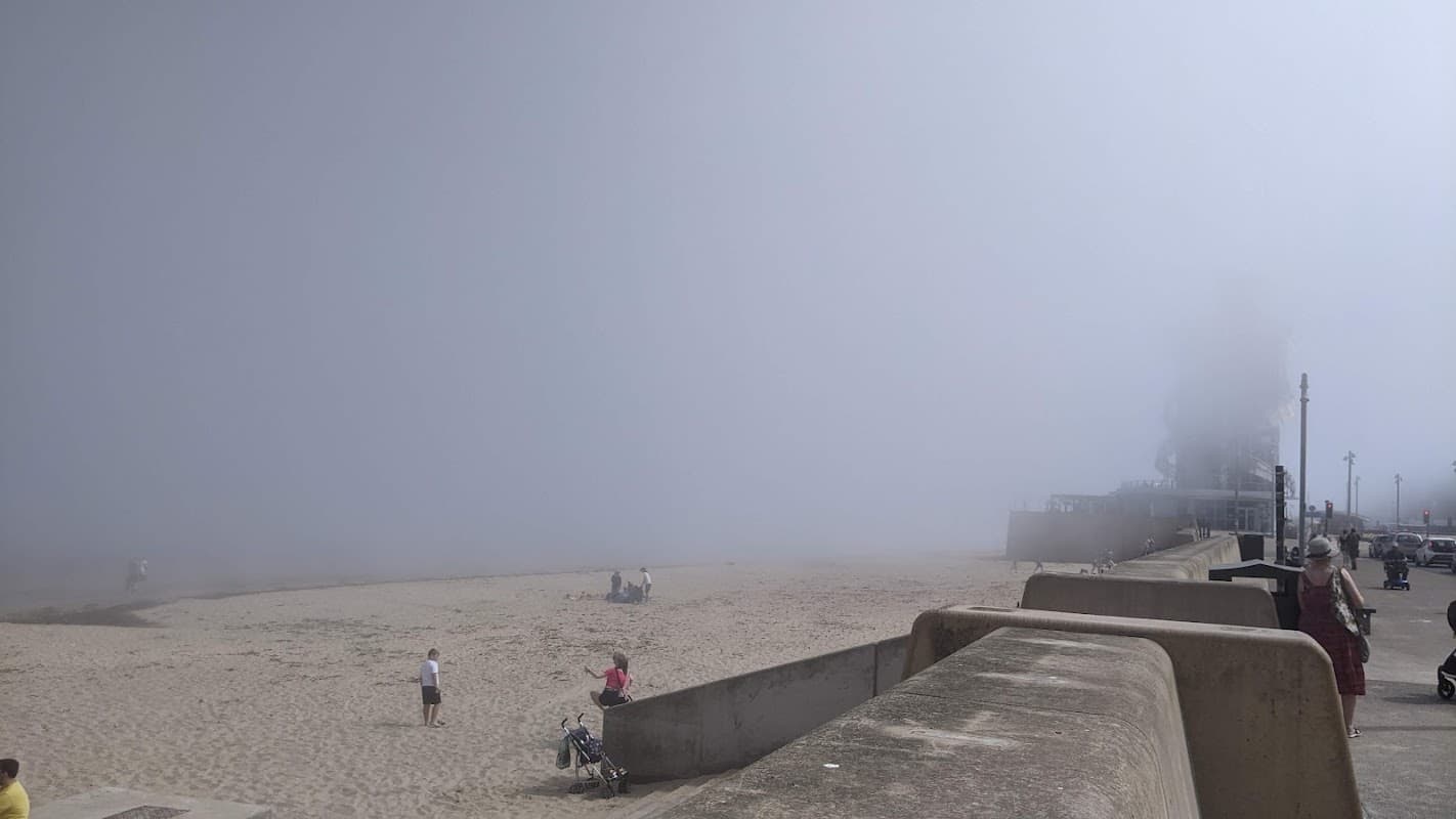 Foggy scene at Turner Street Car Park, with a sandy beach and people near the water's edge.