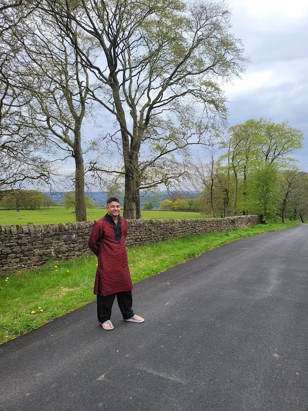 A man in traditional attire stands by a stone wall along a tree-lined road in a lush green landscape.