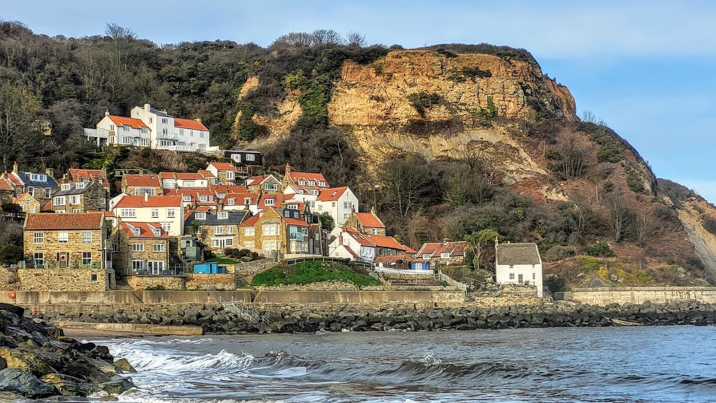 Quaint coastal village with stone houses, cliffs, and a rocky shoreline under a clear blue sky.