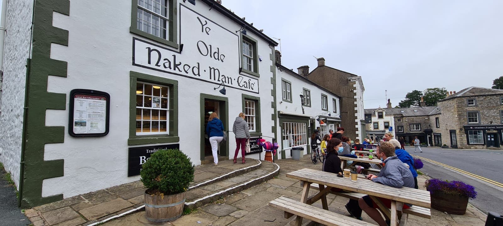 Historic building with "Ye Olde Naked Man CafΓ©" sign, outdoor seating, and people enjoying drinks in Settle, Yorkshire.