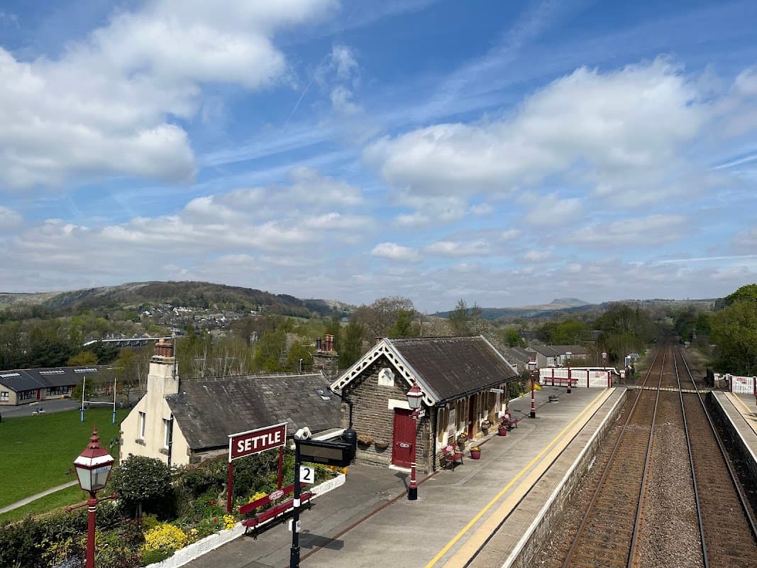 Settle train station with a platform, vintage-style building, and scenic hills under a partly cloudy sky.